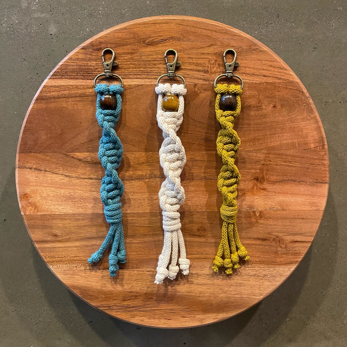 3 Key Chains centered on medium tone wood riser. Twisted style in 3 colors: Teal, Natural White, Spicy Mustard.